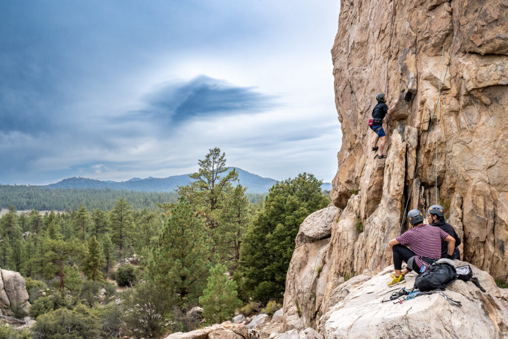 Two Titan Outdoors participants sit on the edge of a rock and watch as a third participant climbs up a large rock that oversees a forest.