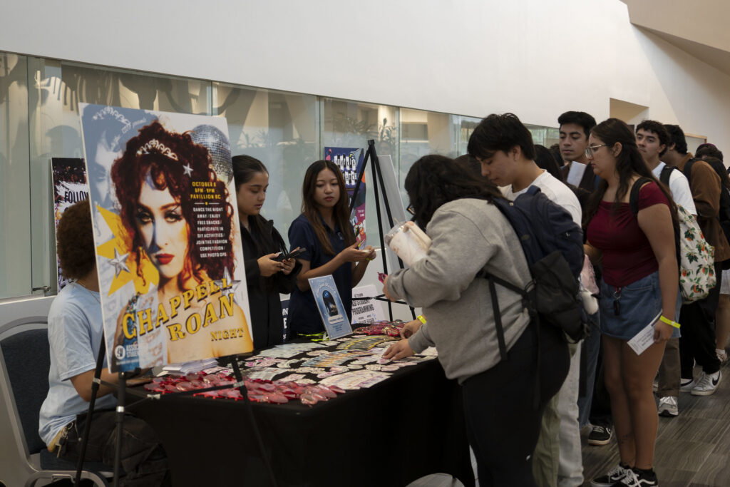 a line of students in front of a Chappell Roan poster and a table filled with Chappell Roan Stickers, handouts, and pink heart glasses.