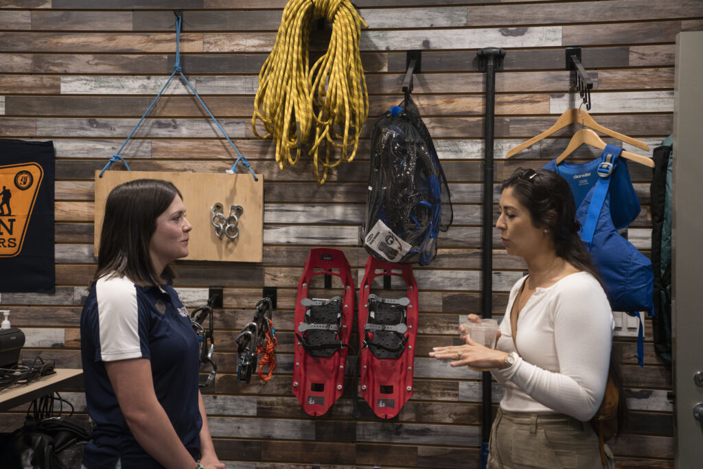 Two young women talk to each other with outdoor rental equipment in the background. 