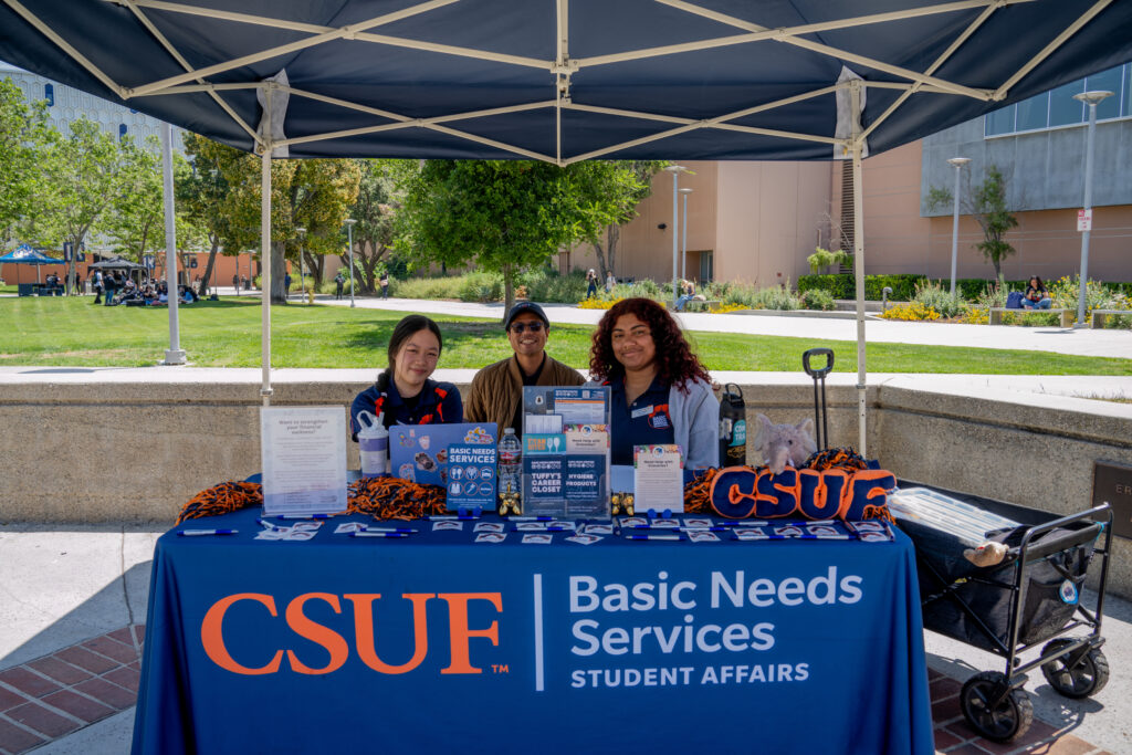 Three people smiling while sat in front of a table that reads "CSUF Basic Needs Services"