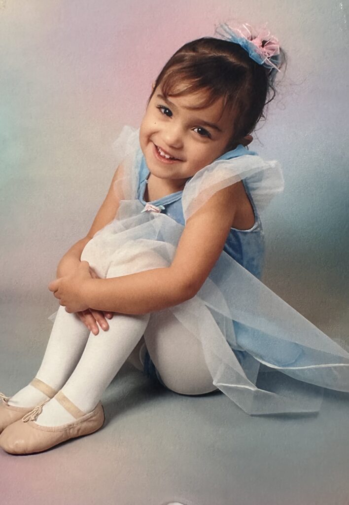 Ashley Mayes as a little kid wearing a blue dress and ballet shoes.