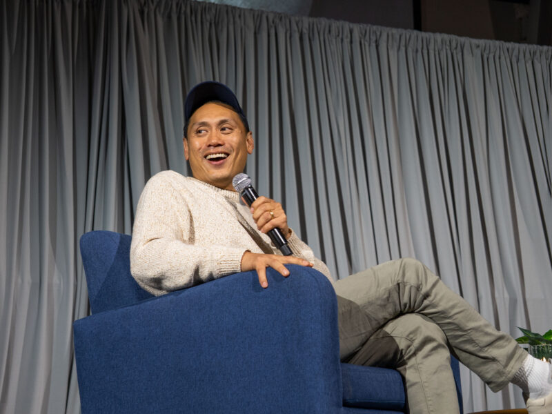 Jon M. Chu smiles at audience on stage with microphone.