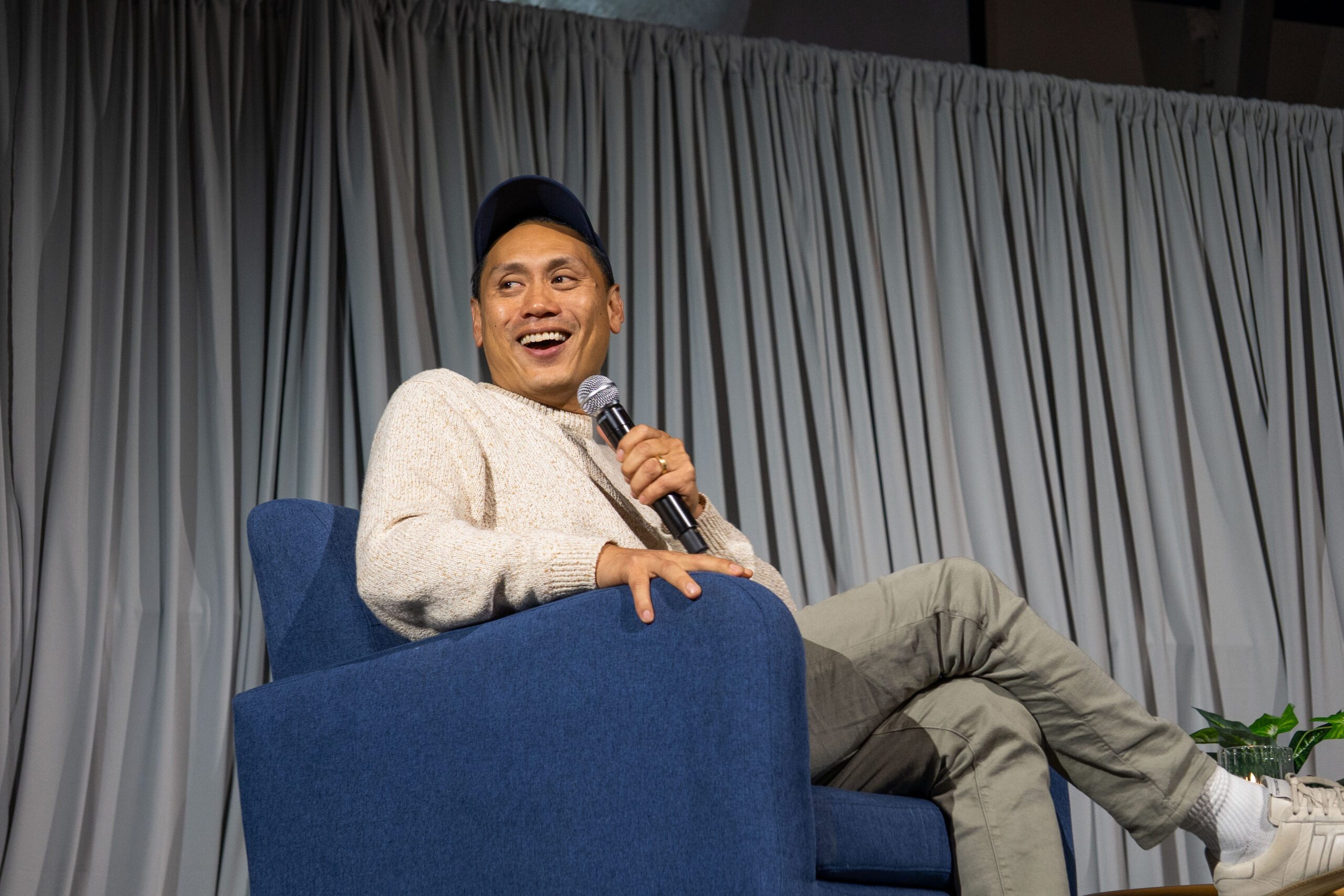 Jon M. Chu smiles at audience on stage with microphone.