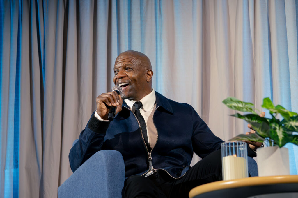 A man, Terry Crews sits in a chair holding a microphone looking out on the audience.