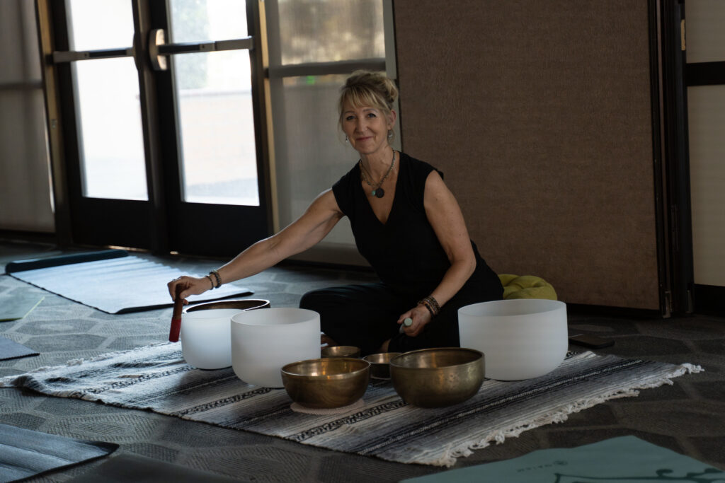 A woman holding a stick sits around metal sound bowls.