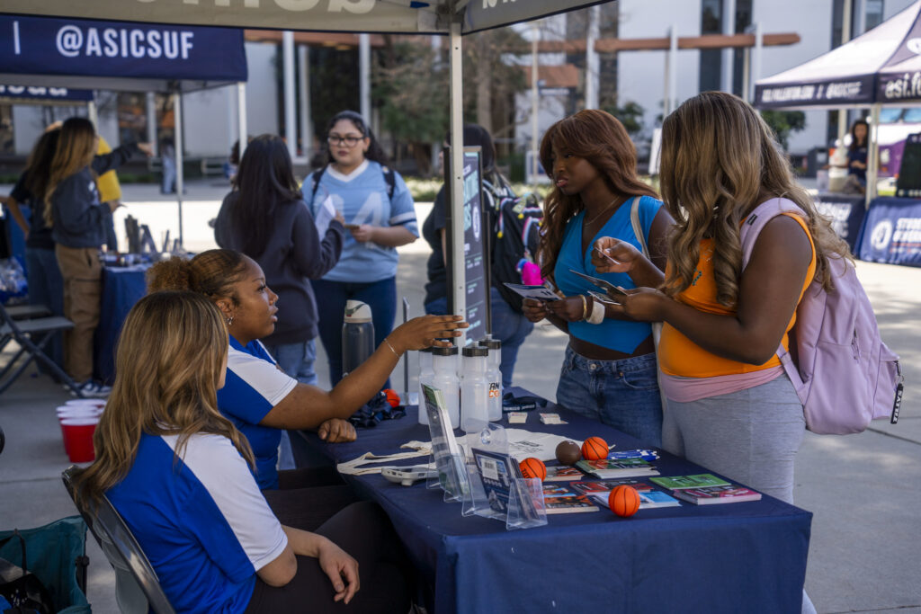 Two girls sit on one side of a table while one gestures to two girls standing on the other side.