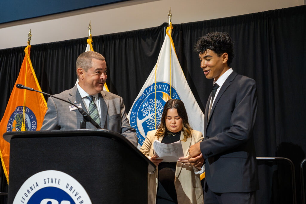 two men have a conversation on a stage with a woman in the background 