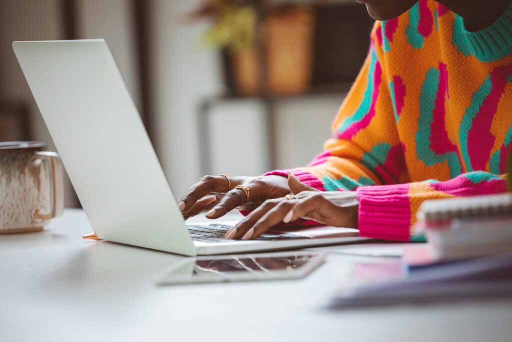 Afro american young woman wearing colorful sweater sitting at the desk at home and using laptop