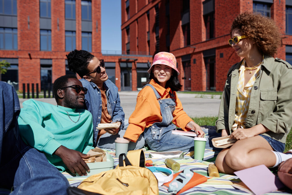 Diverse group of cheerful young students enjoying picnic outdoors on grass in modern college campus, focus on Asian young woman smiling